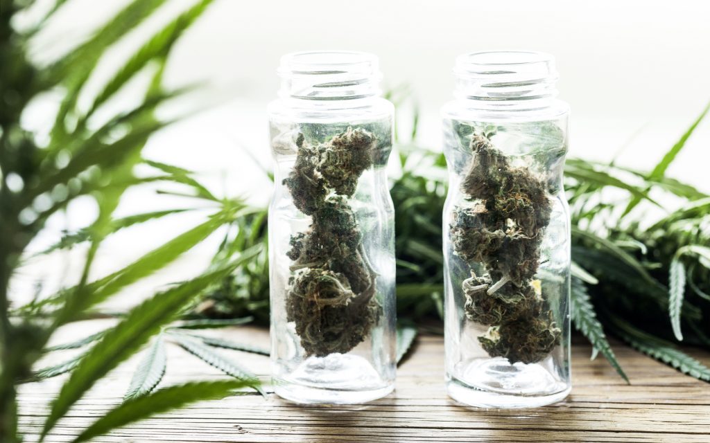 Marijuana buds in small jars on wooden table with cannabis leaves