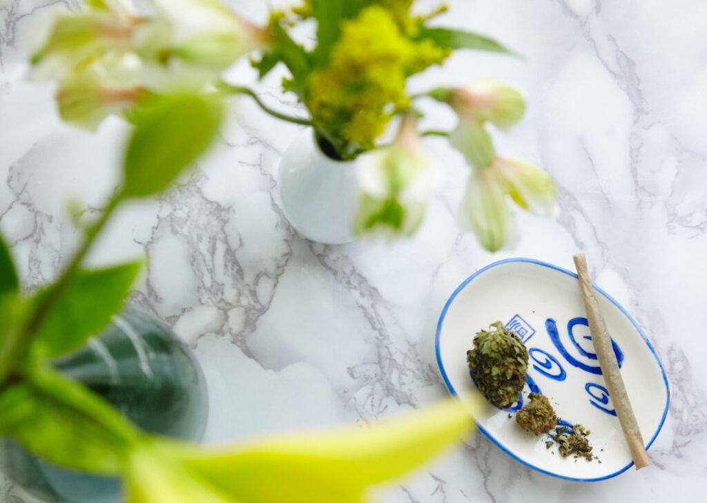 A high angle shot of a marijuana blunt and pieces of dry cannabis with blurred flowers in vases