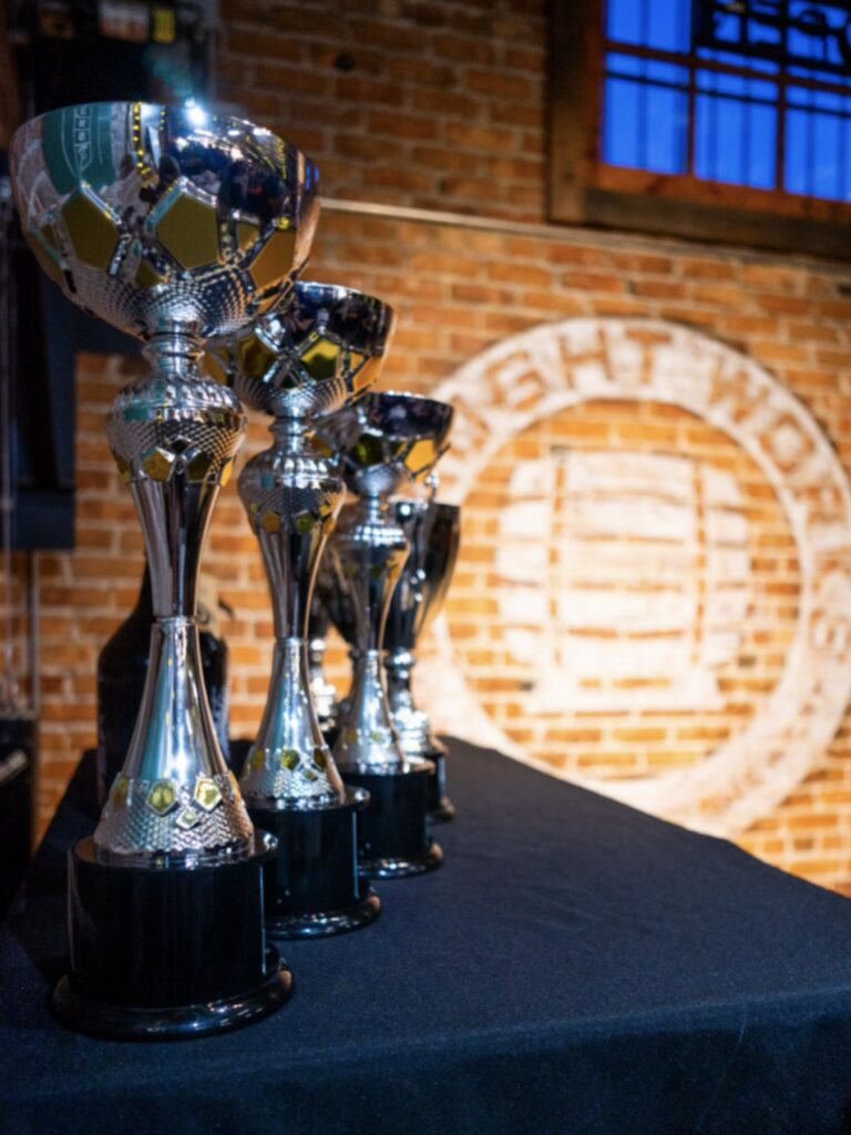 four large gleaming trophies stand on a table at Draught Works brewery in front of an exposed brick wall and a window revealing dusk light