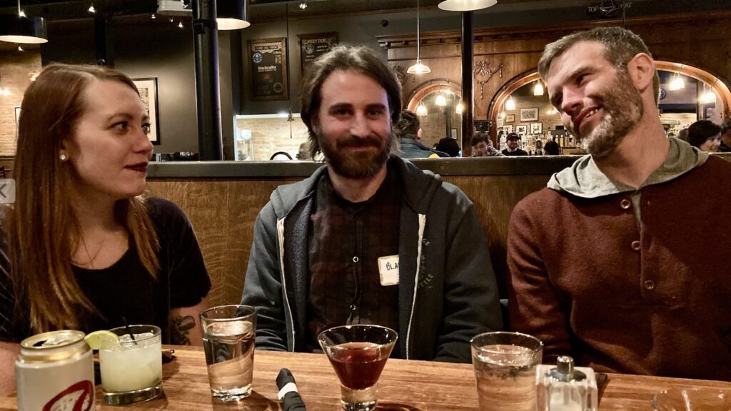 a woman and two men wearing relieved smiles drink cocktails at a wood paneled bar in Missoula