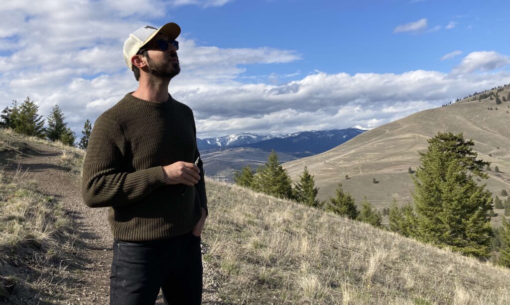 man in black jeans and green sweater stands on a rise holding a vape pen with a snowy peak in the background