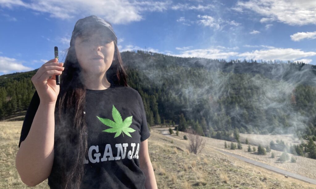 young woman with long brown hair and black t shirt reading Laganja with a green pot leaf exhales from a vape pen in front of a large stand of pine trees